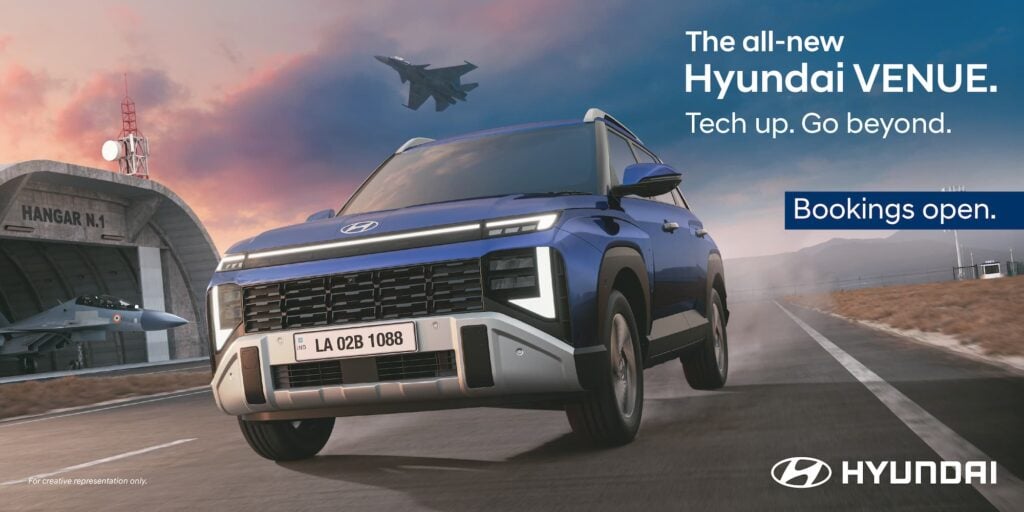 Dynamic front view of the all-new 2025 Hyundai Venue in Mystic Sapphire blue, driving on a road near an aircraft hangar."