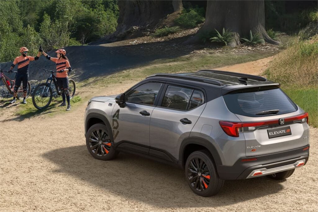 Rear three-quarter view of the dual-tone Honda Elevate ADV (silver/grey and black) on a dirt trail, featuring large 'ADV' side decals and black alloy wheels with orange accents. Two cyclists are standing nearby.